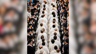 A procession of people in white robes walks down an aisle flanked by uniformed individuals. Crowd watches from sides, creating a solemn mood.
