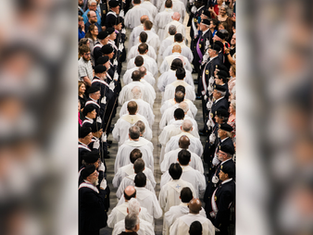 A procession of people in white robes walks down an aisle flanked by uniformed individuals. Crowd watches from sides, creating a solemn mood.