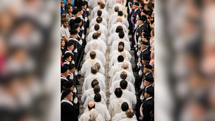 A procession of people in white robes walks down an aisle flanked by uniformed individuals. Crowd watches from sides, creating a solemn mood.