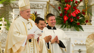 Clergymen in white, smiling and clapping, stand in an ornate church with red flowers in the background. One holds a paper. Joyful mood.