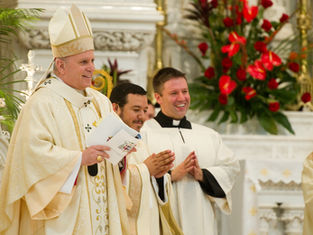 Clergymen in white, smiling and clapping, stand in an ornate church with red flowers in the background. One holds a paper. Joyful mood.