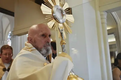 A person holds a large golden monstrance in a church setting, wearing a white robe. Others are present in the background. The mood is solemn.