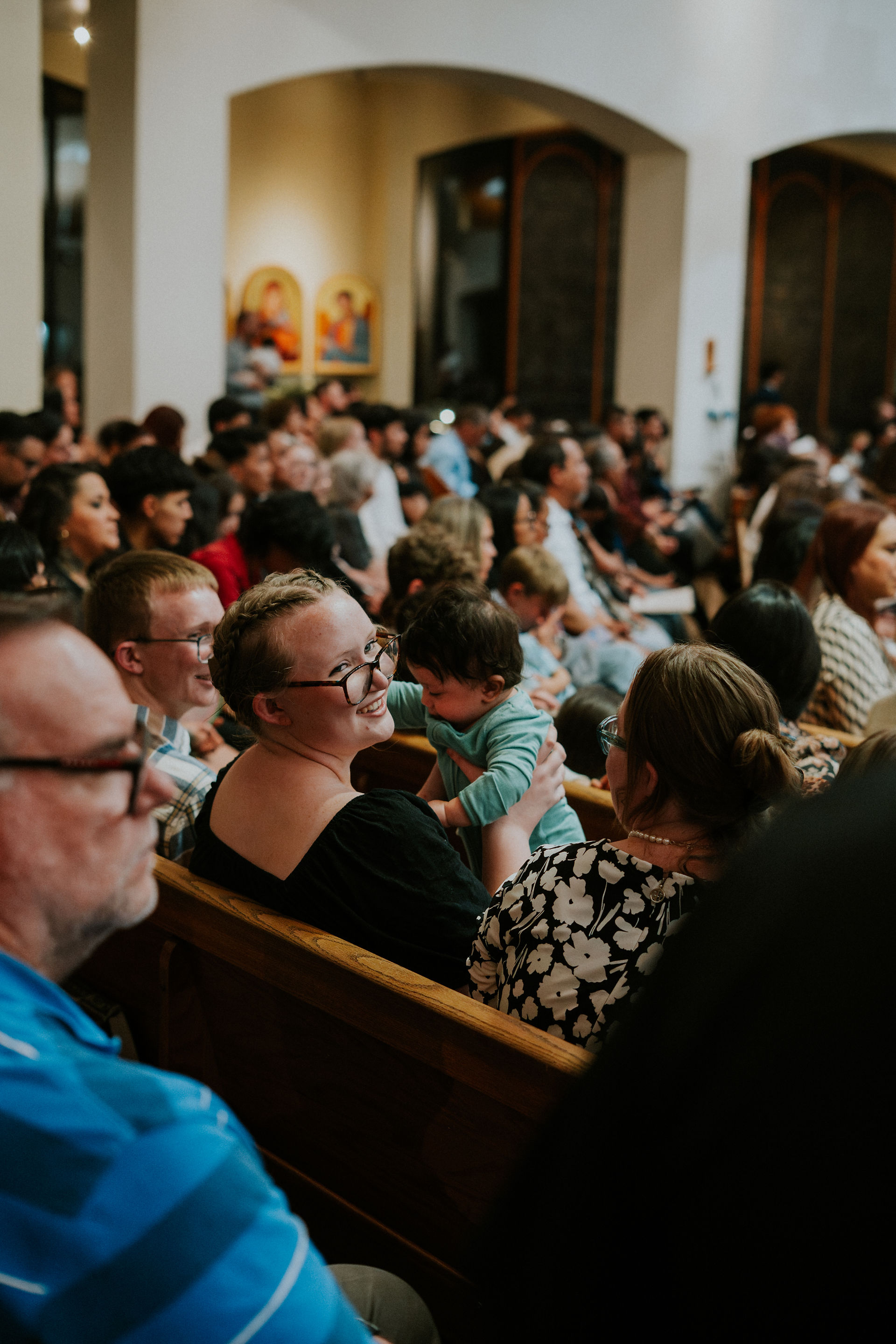 Crowded gathering in a hall. A woman smiles, holding a baby in teal. Attendees sit on wooden benches, focusing ahead. Wall art visible.