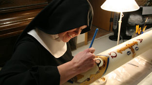 A nun in black habit paints religious symbols on a large candle in a warmly lit room, smiling as she works under a table lamp.