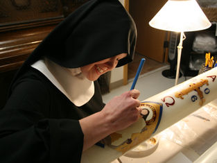 A nun in black habit paints religious symbols on a large candle in a warmly lit room, smiling as she works under a table lamp.