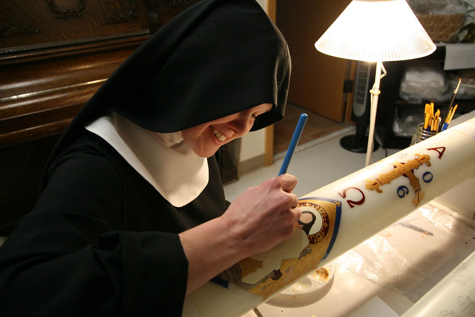 A nun in black habit paints religious symbols on a large candle in a warmly lit room, smiling as she works under a table lamp.