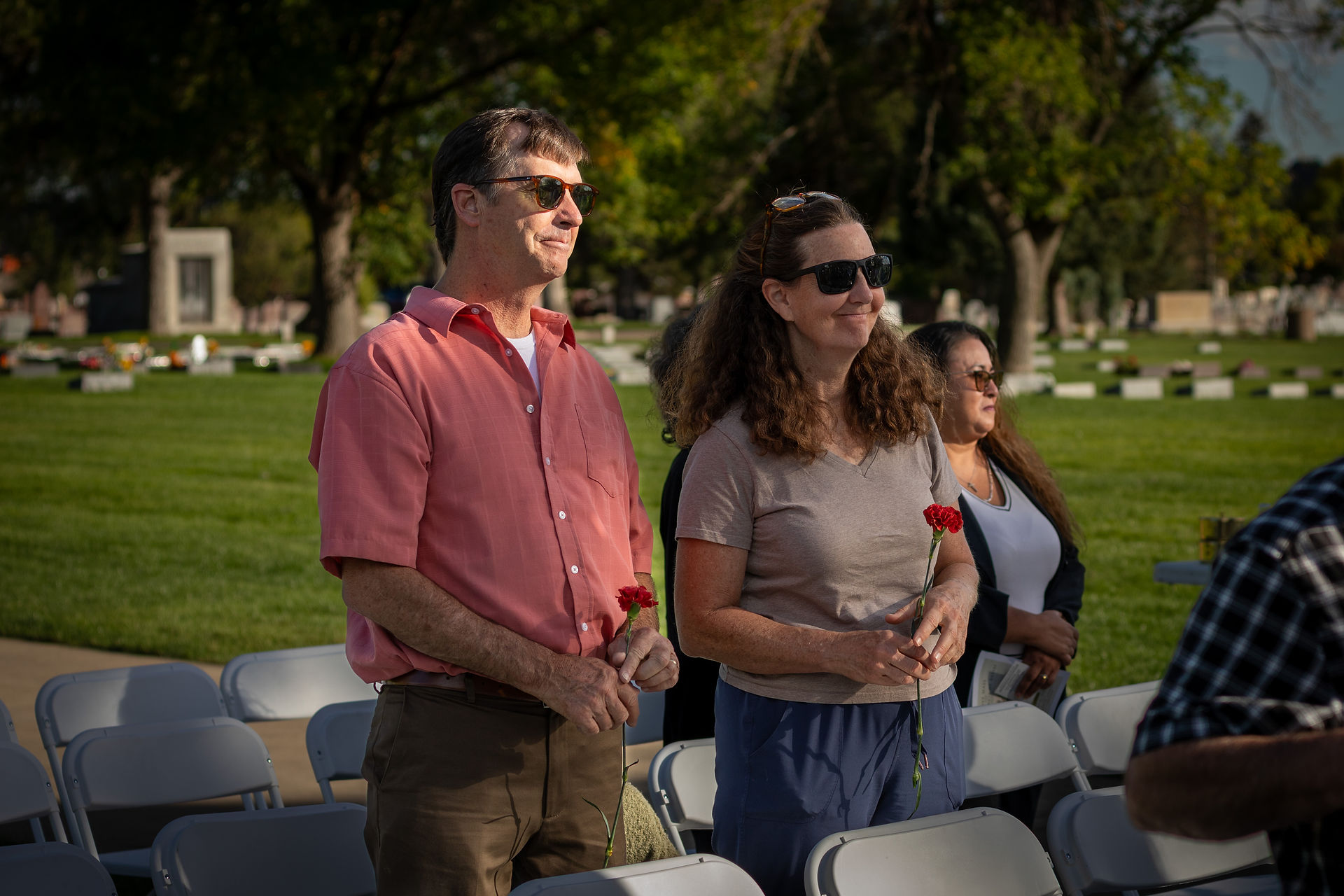 Two people holding red carnations stand in a grassy cemetery under a blue sky, wearing sunglasses, surrounded by empty chairs.