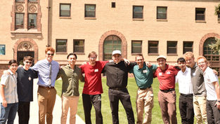 A group of eleven smiling men stand arm in arm on grass, with a brick building in the background. Bright, sunny day with casual attire.