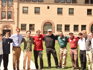 A group of eleven smiling men stand arm in arm on grass, with a brick building in the background. Bright, sunny day with casual attire.