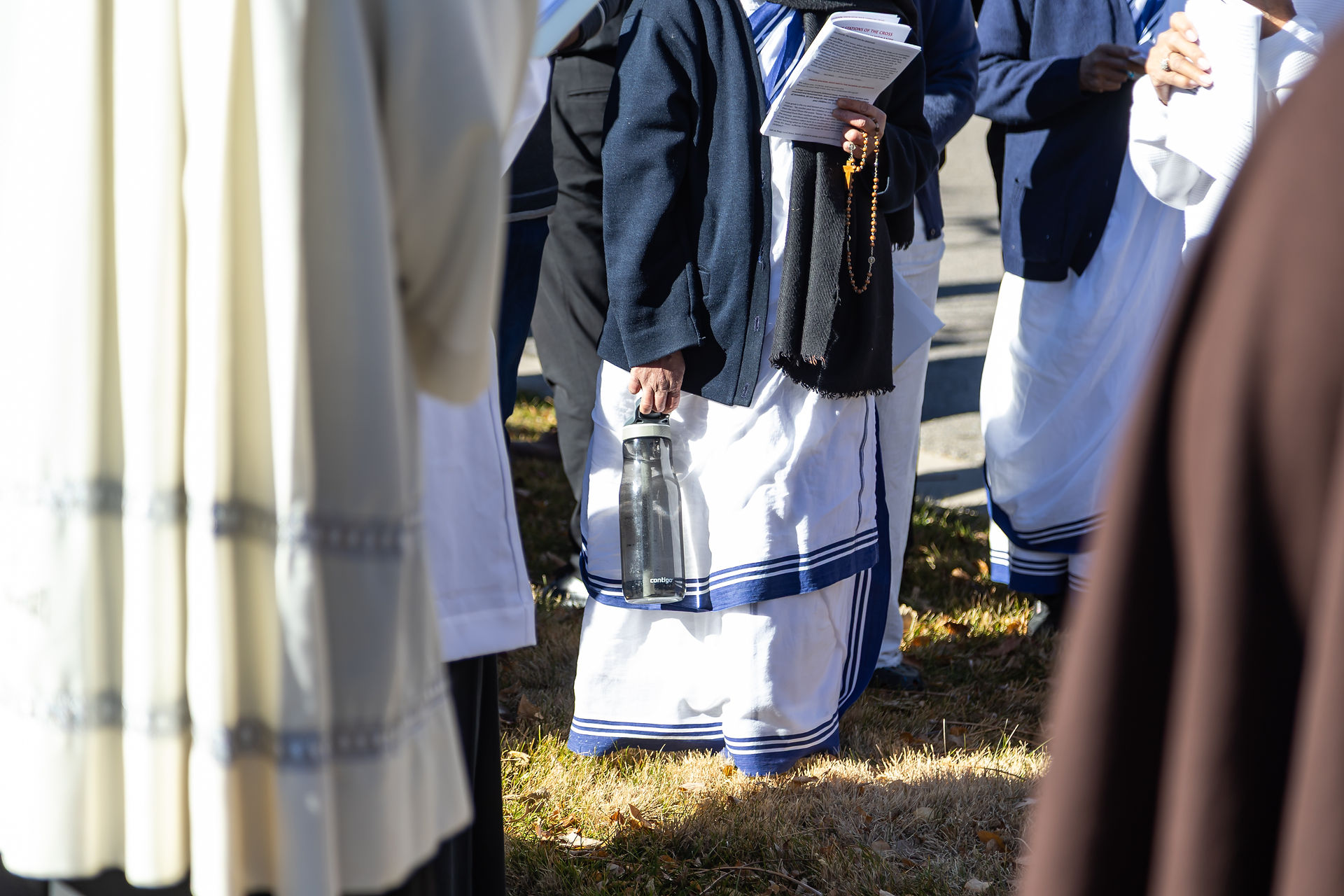 People in religious garments hold papers and a rosary, one carrying a water bottle. They're outdoors on grass, sunlight casting shadows.