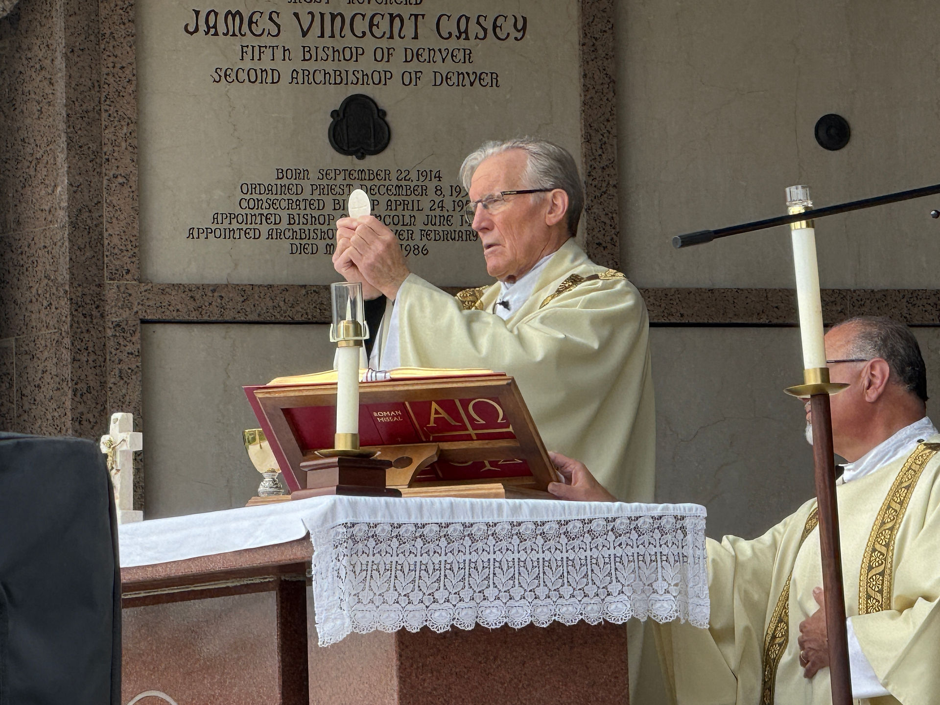 Priests in cream vestments hold a service at an altar with a lace cloth, candles, and red book. Stone wall with engraved text in background.