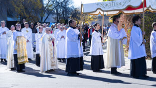 A religious procession with people in white robes under a decorative canopy. Trees with autumn leaves and a crowd are visible in the background.