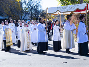 A religious procession with people in white robes under a decorative canopy. Trees with autumn leaves and a crowd are visible in the background.