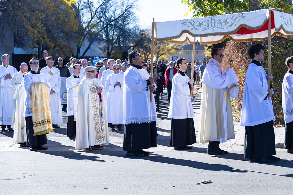 A religious procession with people in white robes under a decorative canopy. Trees with autumn leaves and a crowd are visible in the background.