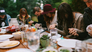 People sitting at a table outdoors, holding hands in prayer. Plates and glasses present, soft focus, greenery in the background.