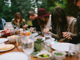 People sitting at a table outdoors, holding hands in prayer. Plates and glasses present, soft focus, greenery in the background.
