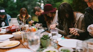 People sitting at a table outdoors, holding hands in prayer. Plates and glasses present, soft focus, greenery in the background.