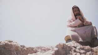 Person in a beige robe sits on rocky terrain under a clear sky, hands clasped in thought. Peaceful and contemplative mood.