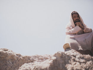 Person in a beige robe sits on rocky terrain under a clear sky, hands clasped in thought. Peaceful and contemplative mood.