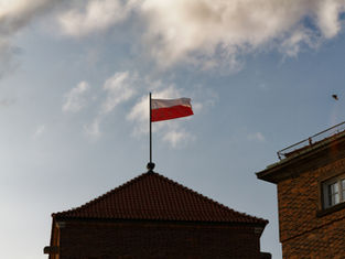 Polish flag on a building rooftop against a blue sky with scattered clouds; a bird flies in the distance. Calm, clear day.