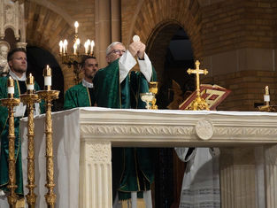 Priests in green vestments celebrate Mass at an ornate altar with candles, a gold cross, and a large book in a dimly lit church.