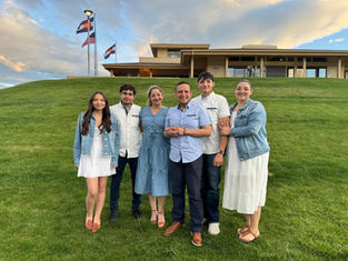A group of six people stands smiling on a grassy hill, with a building and flagpoles in the background under a partly cloudy sky.