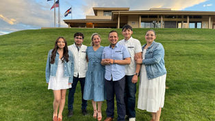 A group of six people stands smiling on a grassy hill, with a building and flagpoles in the background under a partly cloudy sky.