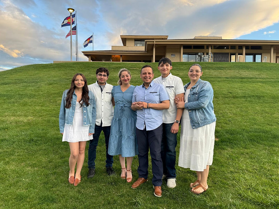A group of six people stands smiling on a grassy hill, with a building and flagpoles in the background under a partly cloudy sky.