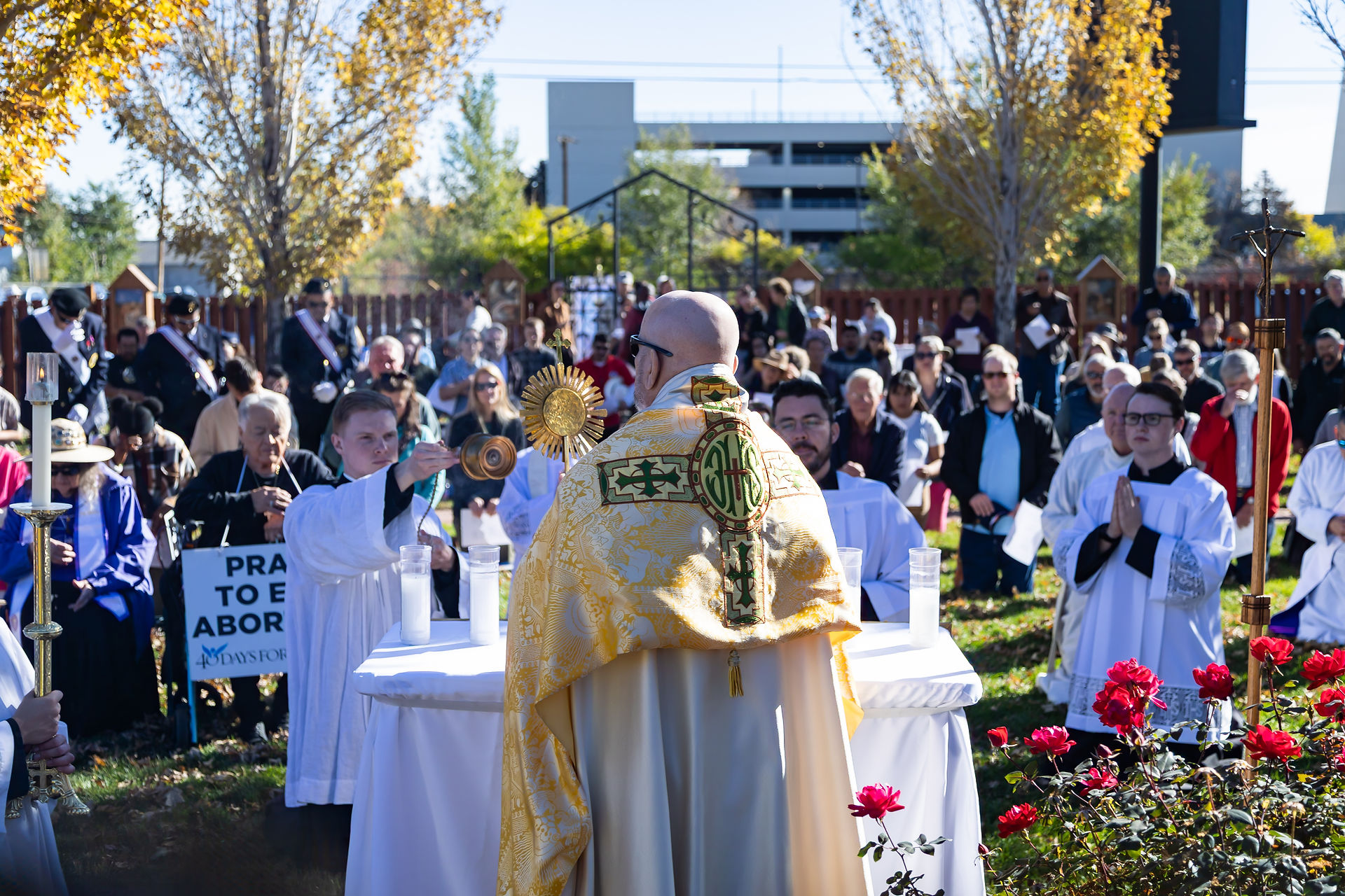 Archbishop in golden vestment leads outdoor religious ceremony. Congregation kneels on grass. "Pray to End Abortion" sign visible.