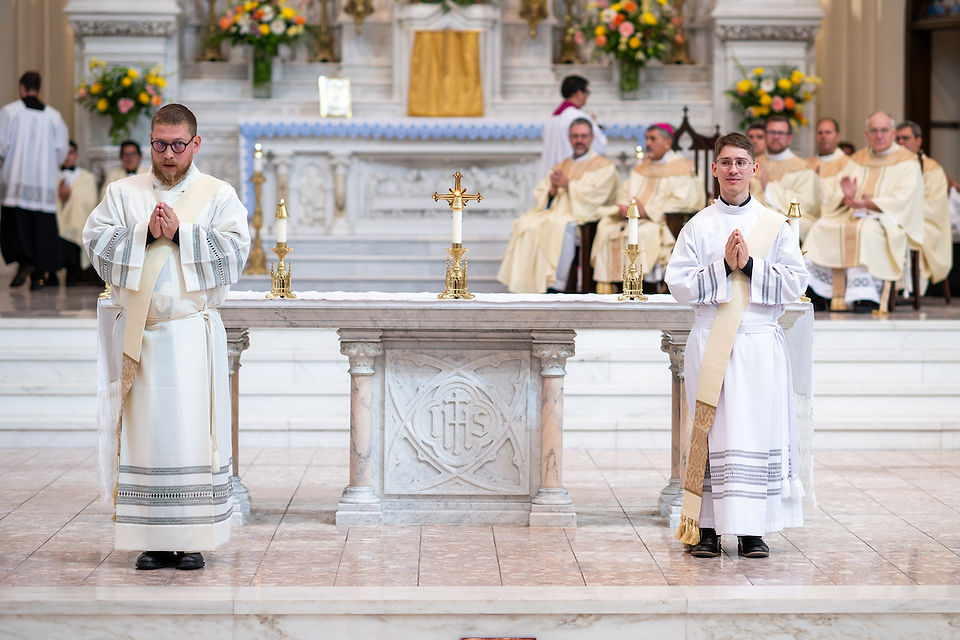 Two clergy in white robes stand in prayer behind a marble altar, with candles and a cross. Background shows seated clergy and flowers.