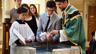 Priest baptizes a baby at a stone font while family and altar boys watch in a church, solemn and warm-lit.