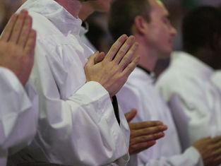 People in white robes stand with hands clasped in prayer. The setting is a solemn indoor gathering with soft lighting, creating a serene mood.