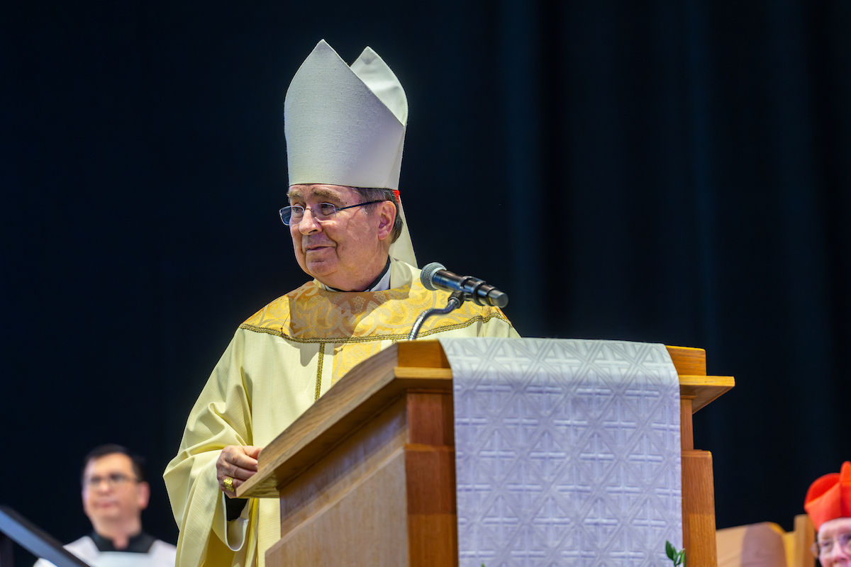 A bishop in white and gold robes stands at a podium speaking into a microphone. A priest is in the background. The scene is formal.