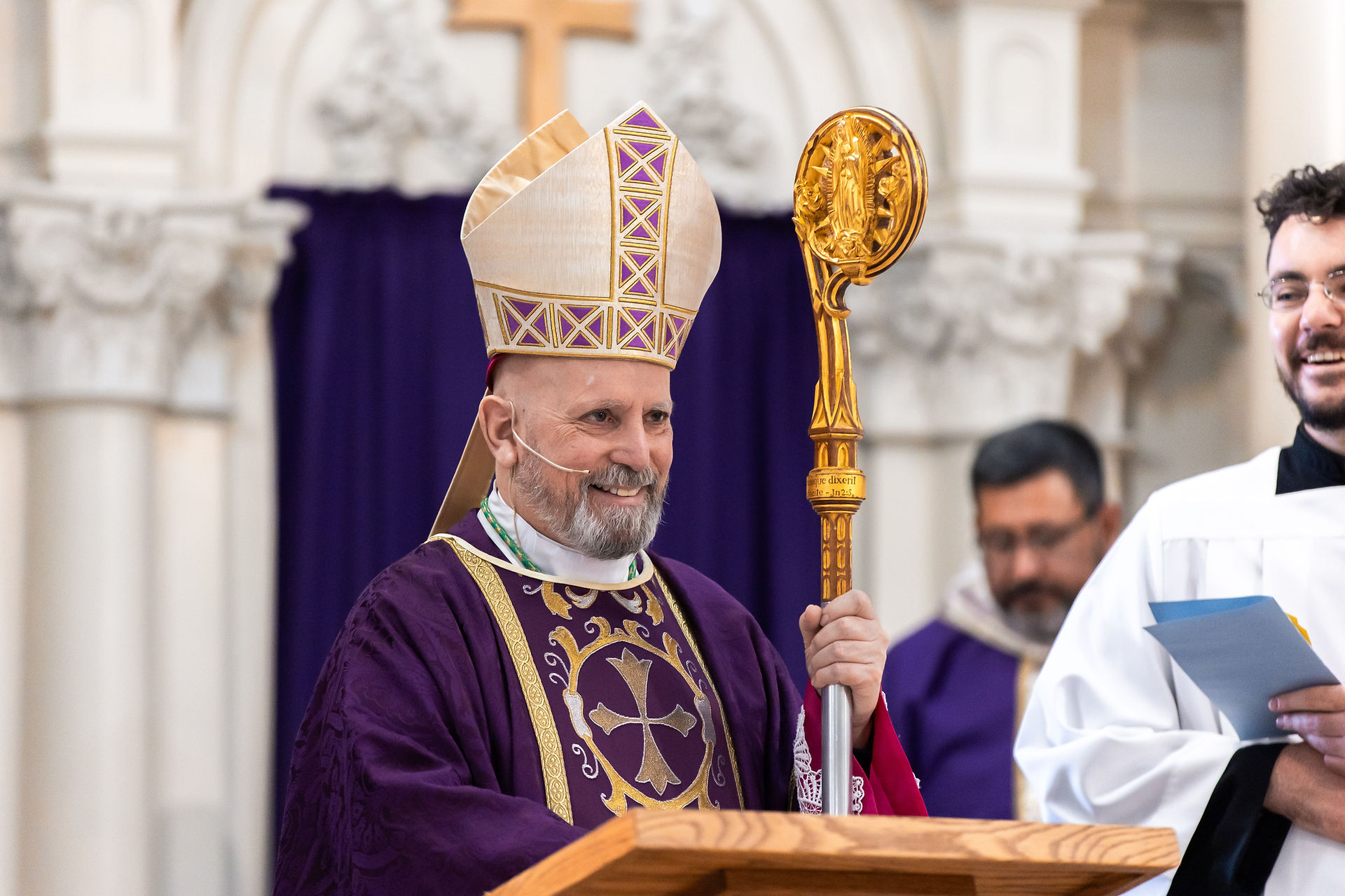 A bishop in purple vestments and mitre holds a gold crosier, smiling during a church service. Background features ornate white pillars.
