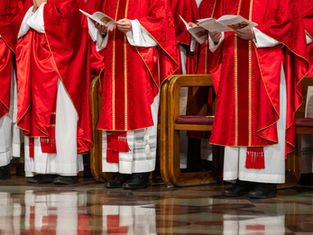 Clergy members in red robes hold service programs, standing on a polished floor with wooden chairs behind them. The mood is solemn.