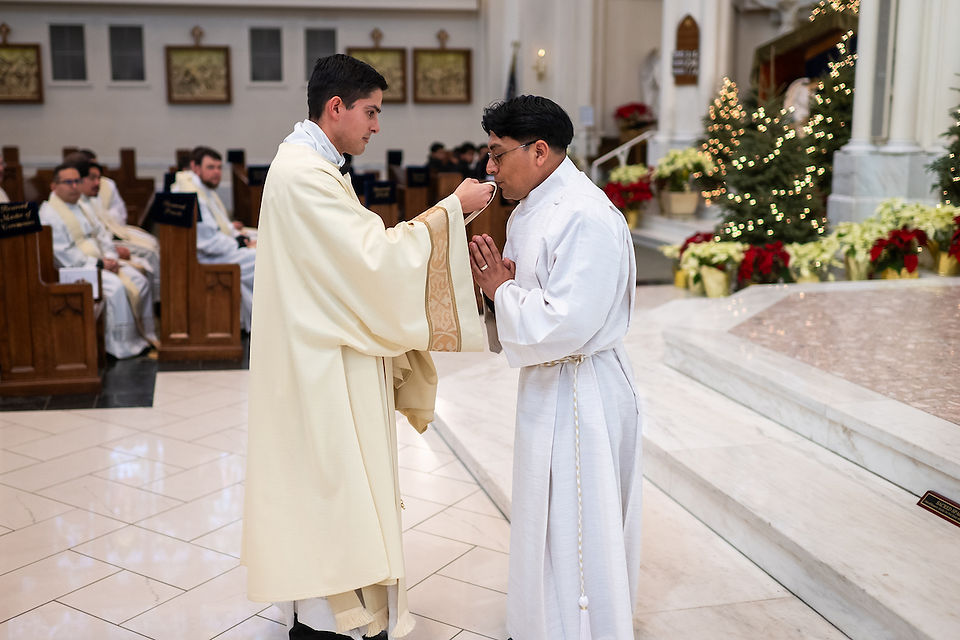 Deacon in white robe offers a religious stole to man in church, who kisses the stole. Pews with attendees in background, Christmas trees with lights, festive decor.