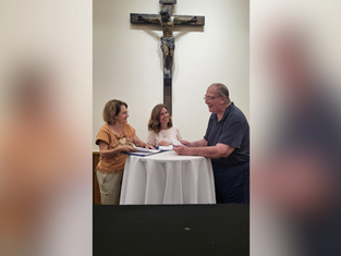 Three people smiling and talking around a round table in front of a crucifix, with documents in hand. Warm lighting and casual attire.