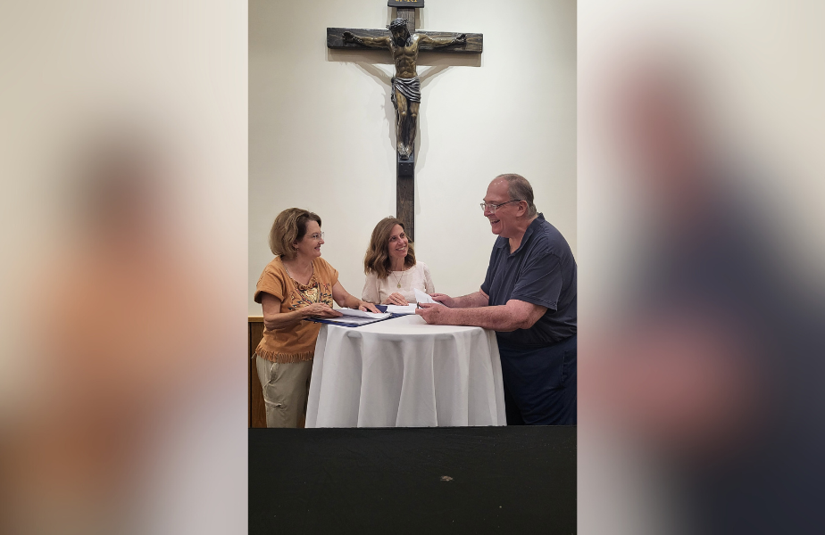 Three people smiling and talking around a round table in front of a crucifix, with documents in hand. Warm lighting and casual attire.