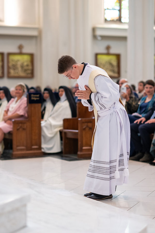 A man in a white robe bows in a church, surrounded by seated nuns and attendees. Bright interior, serene atmosphere.