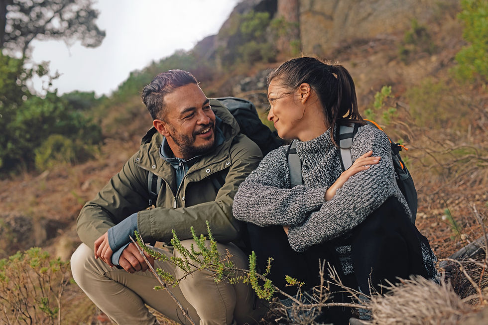 A couple sits, looking at each other, in long grass during a hike. They're carrying backpacks. The mood is happy.