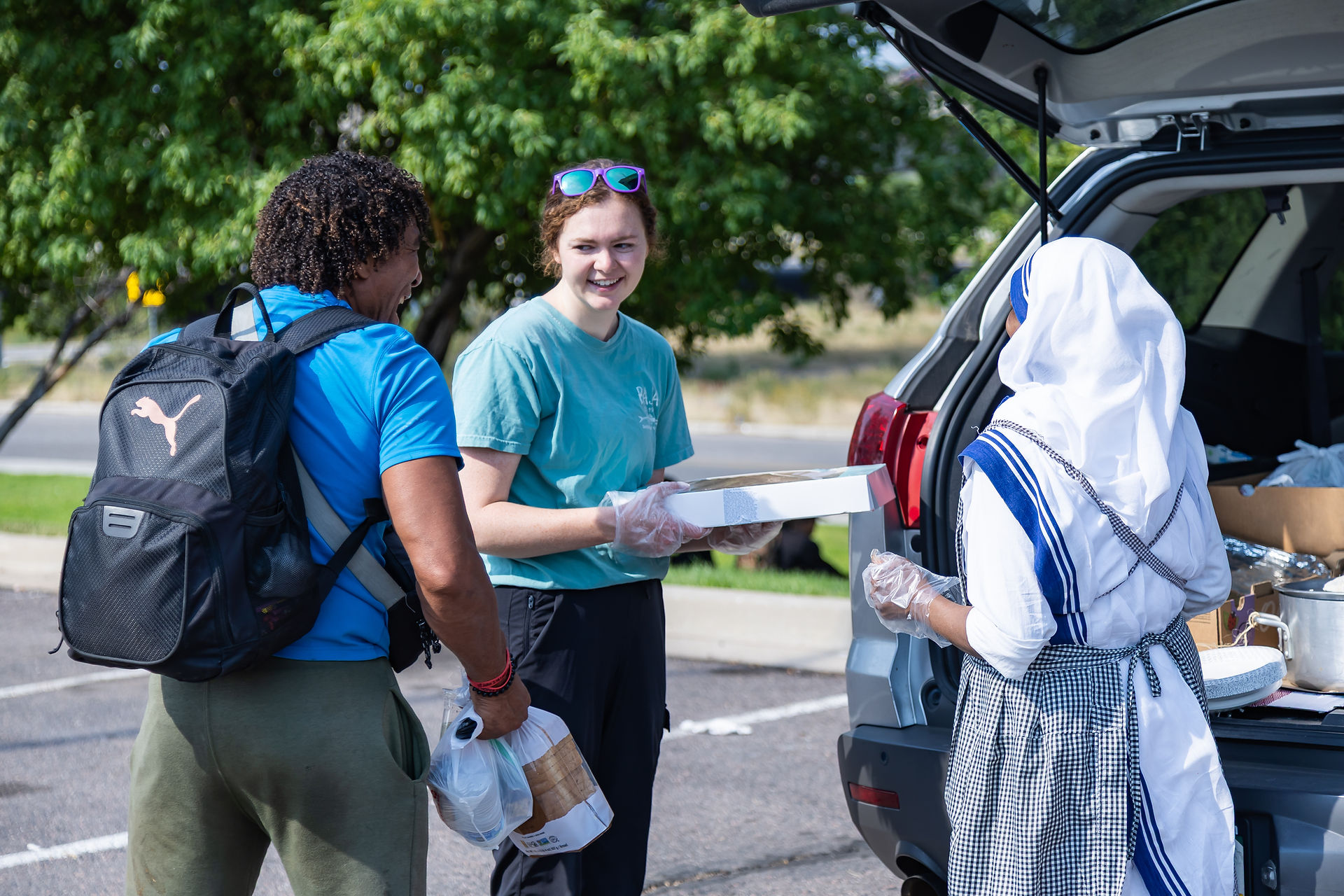 Three people distribute food from a car trunk outdoors. One holds a box of donuts. Green trees in the background create a cheerful setting.