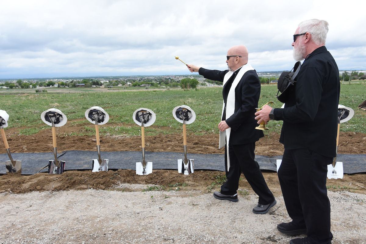 Two men in black suits conduct a ceremonial groundbreaking with shovels and hard hats. Cloudy sky over a grassy field in the background.