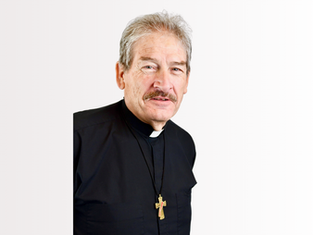 Deacon in black robe with a gold cross necklace. He has grey hair and a mustache, standing against a plain white background, expressing calmness.
