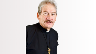 Deacon in black robe with a gold cross necklace. He has grey hair and a mustache, standing against a plain white background, expressing calmness.