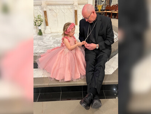 A girl in a pink dress and floral headband sits with a man in black attire on church steps, holding hands. Marble decor in background.