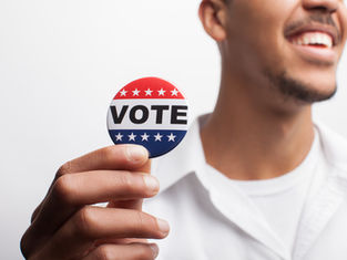 Smiling man holds a red, white and blue VOTE button with stars against a plain white background.