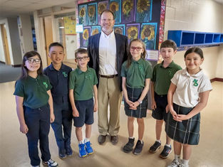 A teacher poses with six students in a school hallway. They wear green and white uniforms, standing in front of colorful sunflower artwork, smiling.