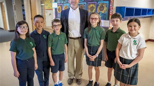 A teacher poses with six students in a school hallway. They wear green and white uniforms, standing in front of colorful sunflower artwork, smiling.