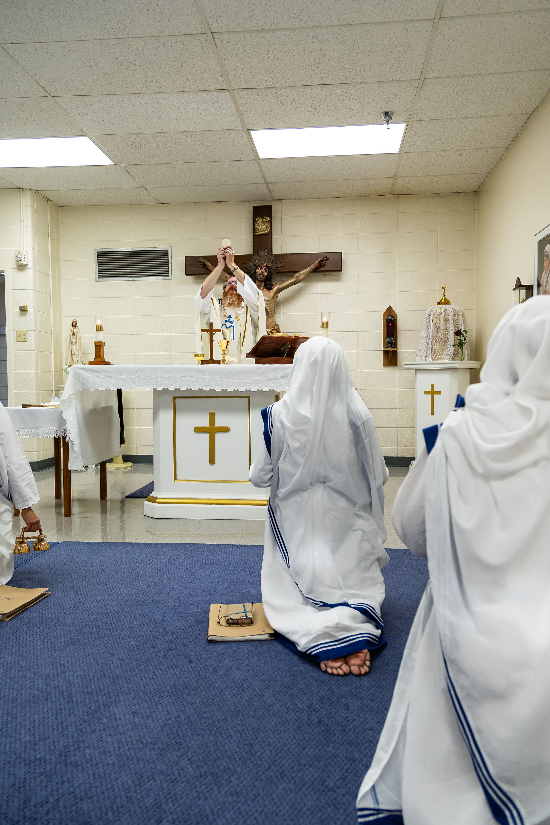 Priest in white robe holds up Eucharist in a chapel. Nuns in blue-striped habits kneel on a blue carpet, facing the altar. Calm, reverent mood.