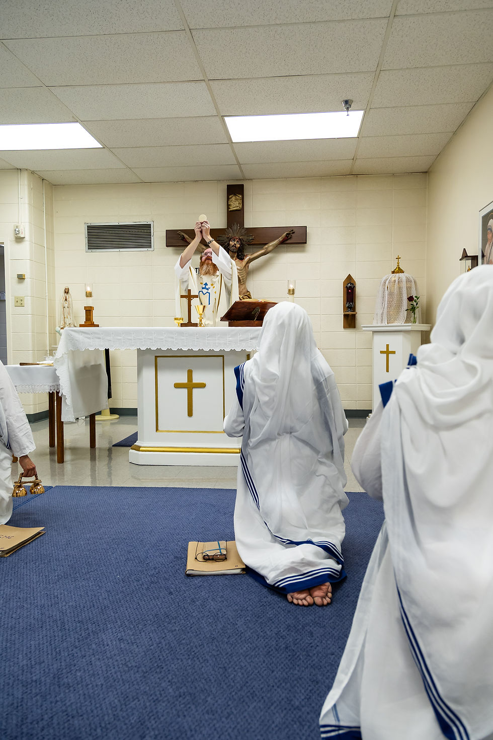 Priest holds up host, nuns kneel in prayer. Church altar with crucifix, candles. White robes with blue stripes, solemn atmosphere.